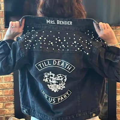 Black denim jacket with white embellishments and text held by a person against a brick wall.