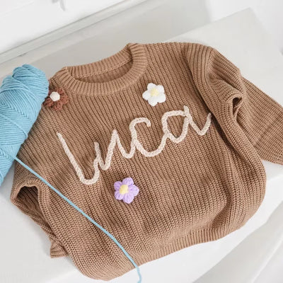 Brown sweater with 'luca' embroidered on it, next to a ball of yarn on a white surface