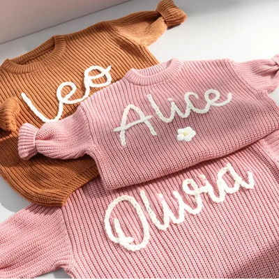 Two pink knitted sweaters with embroidered names on a light background, branded 'Lamoriea'.