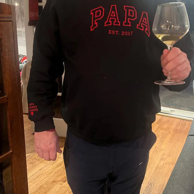 Person wearing a black sweatshirt with 'PAPA EST. 2017' text, holding a glass of white wine indoors.