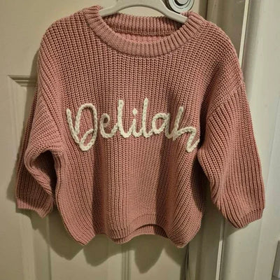 Pink sweater with 'Delilah' text on a hanger against a white door.