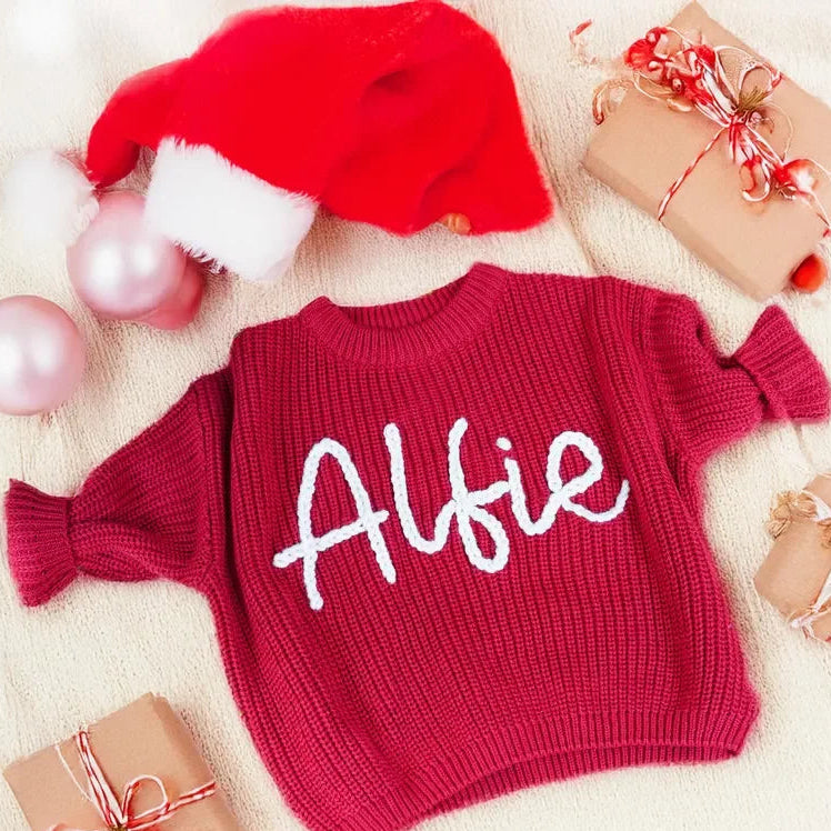 Red sweater with 'Alfie' printed on it, surrounded by Christmas decor including a Santa hat and presents.