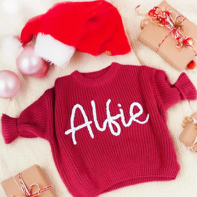 Red sweater with 'Alfie' printed on it, surrounded by Christmas decor including a Santa hat and presents.