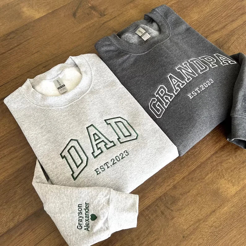 Two folded sweatshirts with 'DAD EST 2023' and 'GRANDPA EST 2023' text on a wooden surface.