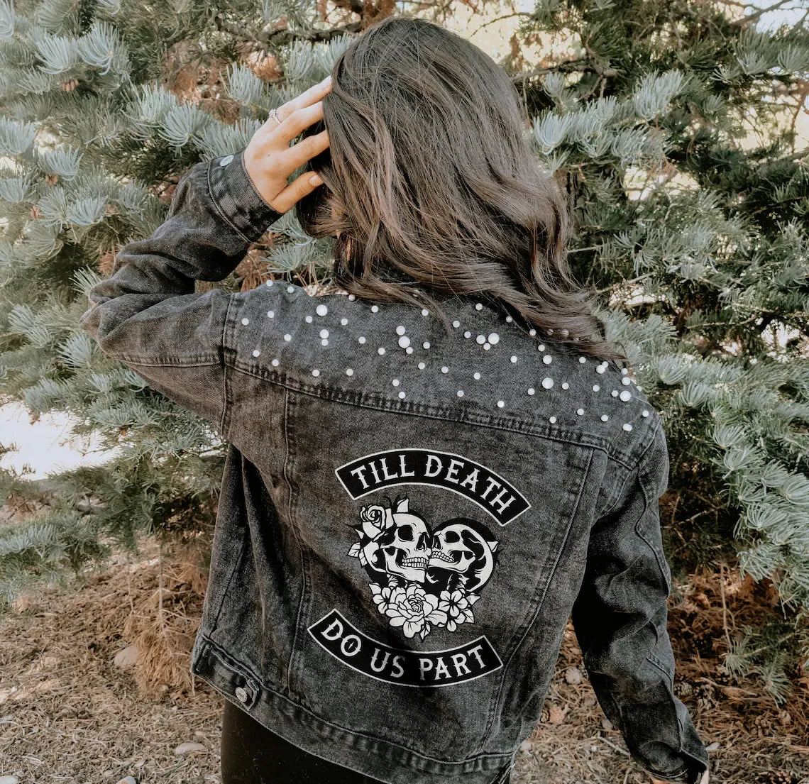Person wearing a denim jacket with decorative skull and rose elements and text, standing in a natural setting.