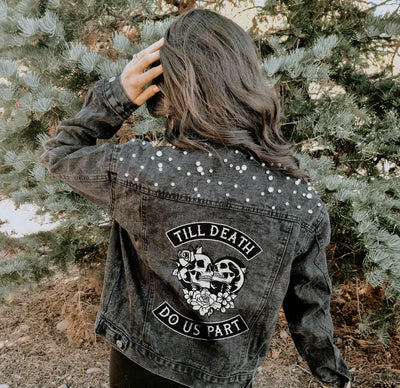 Person wearing a denim jacket with decorative skull and rose elements and text, standing in a natural setting.