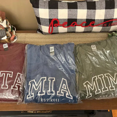 Three t-shirts with text on a wooden surface, one with a checkered pillow in the background.