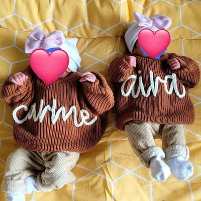 Two baby dolls wearing brown outfits with 'carmine' and 'alina' text, heart-shaped toys, and bows on a yellow patterned blanket.