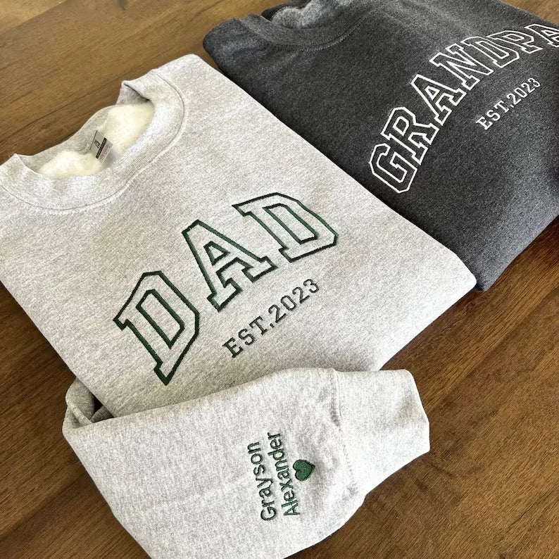 Two folded sweatshirts, one beige with 'MOM EST.1999' text and the other brown with 'DAD' text, on a textured white surface.