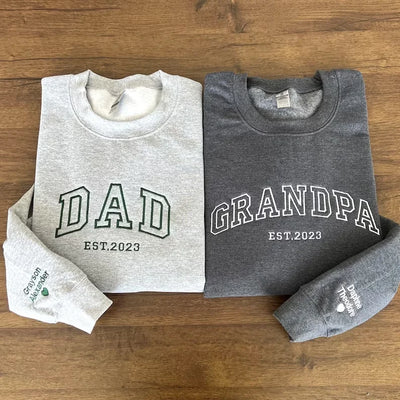 Two sweatshirts, one white and one gray, with 'DAD EST. 2023' and 'GRANDPA EST. 2023' text on a wooden surface.