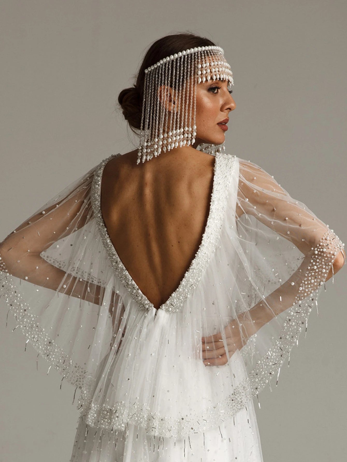 Woman wearing a white wedding dress with a beaded headpiece and sheer sleeves against a neutral background
