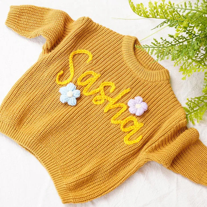 Yellow knitted sweater with 'Sasha' and floral designs on a white background with greenery.